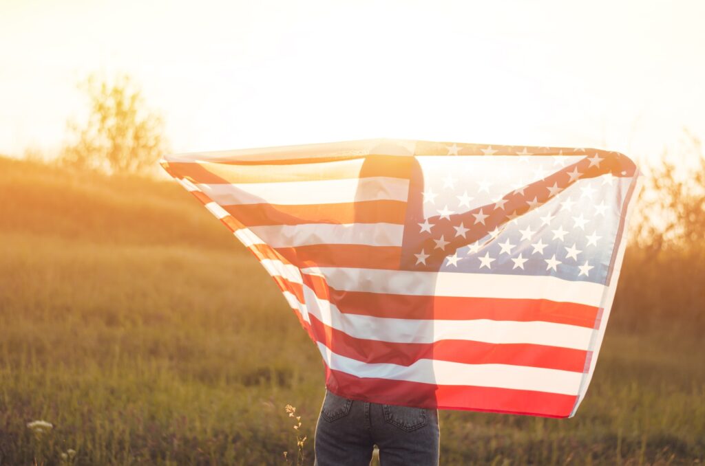 A young woman holds the US national flag in a field at sunset